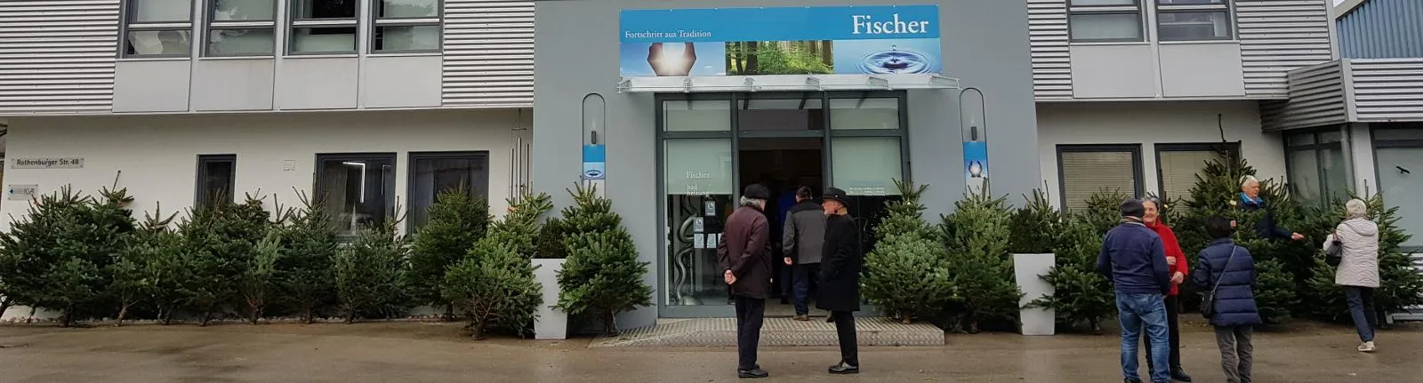 Die Außenansicht des Fischer-Gebäudes zeigt einen modernen Eingangsbereich mit Glasfront, grauer Fassade und einem blauen Firmenschild über der Tür.