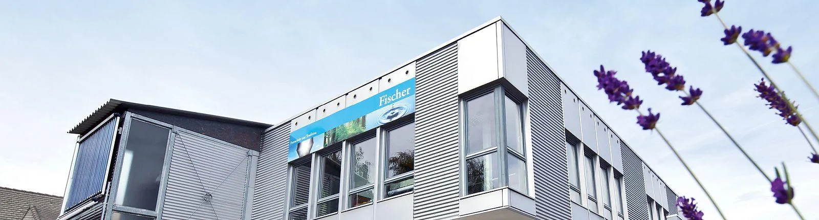 Ein modernes Firmengebäude mit grauer Metallfassade, großen Fenstern und einem blauen Schild mit der Aufschrift „Fischer“ ist aus einer niedrigen Perspektive fotografiert.