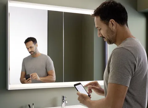 Mann mit Smartphone steht vor einem Badezimmer-Leuchtspiegelschrank der Serie One von Geberit.