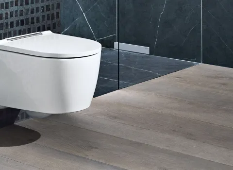 Ein modernes, wandmontiertes WC mit geschlossenem, abgerundetem Kunststoffdeckel. Das Bad hat dunkle Kacheln und einen hellen Holzfußboden.