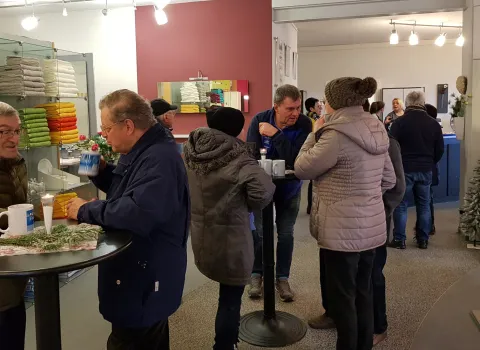 Mehrere Besucherinnen und Besucher stehen in einem hell beleuchteten Ausstellungsraum an Stehtischen zusammen und unterhalten sich bei Getränken. 