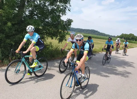 Eine Gruppe von Rennradfahrern fährt bei sonnigem Wetter auf einer ruhigen Landstraße durch eine grüne Landschaft.